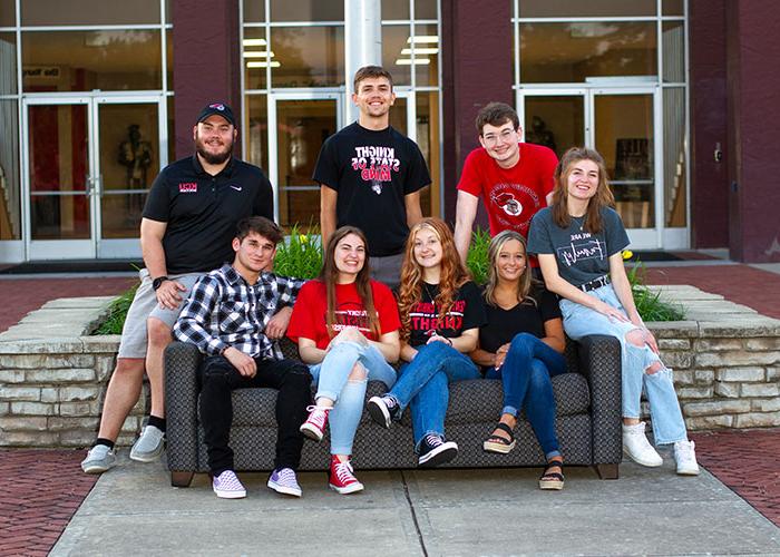 Eight young adults are sitting on and around a couch outdoors, all smiling. They are dressed casually, and a building with large glass windows is visible in the background.